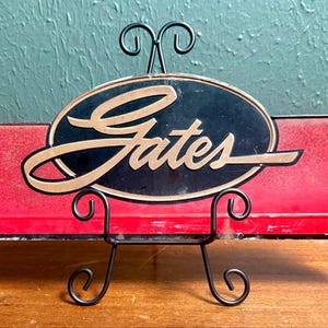 May include: A rectangular red metal sign featuring a central black oval with the word Gates written in gold script lettering. The sign is displayed on a black metal wire stand with decorative scrollwork at the base and top.