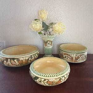 May include: A collection of antique ceramic pieces, including a vase and two bowls, with a decorative relief design. The pieces are in shades of beige, brown, and green, and are arranged on a dark wooden surface. The vase holds a bouquet of yellow flowers.