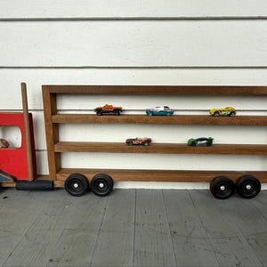 May include: A wooden toy truck display shelf featuring a red and blue cab with a tall smokestack and four black wheels. The attached trailer consists of three horizontal wooden shelves designed to hold small toy cars, with five cars displayed.