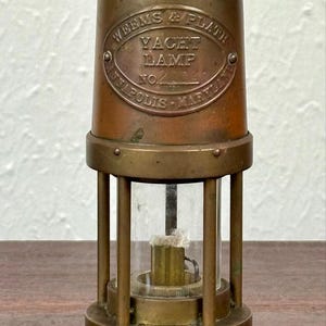 May include: Antique brass yacht lamp with a clear glass cylinder and the text "WEEMS & PLATH YACHT LAMP" on the front. The lamp has a handle on top and is designed with vertical support rods. A vintage nautical item.