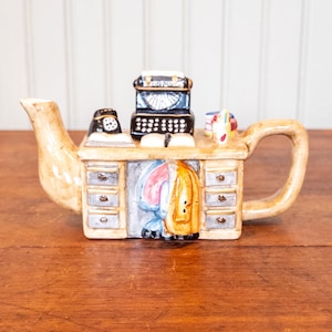 May include: A ceramic teapot shaped like a desk with a tan finish, featuring a black typewriter, a black rotary telephone, and books on the surface. The desk has three drawers on each side and a jacket hanging in the center knee space area.