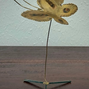 May include: A brass butterfly sculpture with a delicate, openwork design. The butterfly is mounted on a thin wire stem, supported by a three-legged base. The wings have a textured surface and the antennae extend upwards.
