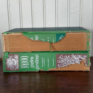 May include: Two vintage green hardcover books titled "10,000 Garden Questions" and "Volume 1" are stacked. The books have green covers with silver text and illustrations. The edges of the books show wear and tear, with exposed wood.