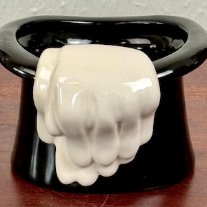 May include: A black ceramic top hat-shaped container with a cream-colored fist-shaped object protruding from the front. The hat has a glossy finish and sits on a wooden surface. The fist is detailed with knuckles and a textured surface.