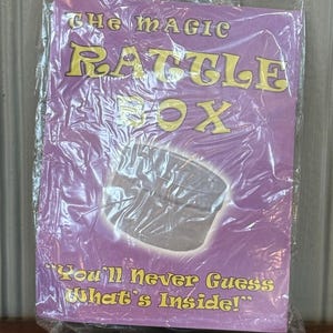May include: A rectangular product package enclosed in clear plastic wrap. The purple packaging features the yellow text THE MAGIC RATTLE BOX at the top and the phrase You'll Never Guess What's Inside! at the bottom, with a central graphic of a box.