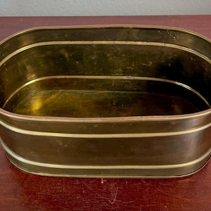 May include: An antique brass planter with an oval shape. The planter has a dark brass exterior with two horizontal white stripes and a bright brass interior. Small handles are on each side.