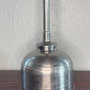 May include: A silver-colored metal oil can with a cylindrical body and a long, narrow vertical spout extending from the center top. The metallic surface shows signs of wear and light scratches, with a smooth, rounded base and a uniform finish.