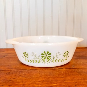 May include: A white milk glass casserole dish with integrated side handles. The exterior features a repeating green floral pattern consisting of stylized daisies and delicate leafy sprigs arranged in a symmetrical horizontal design around the bowl.