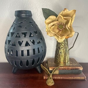 May include: A matte black ceramic lantern with cut-out geometric patterns and three feet, a brass pitcher with embossed figures holding a large artificial magnolia flower, and a small brass eagle figurine resting on two stacked vintage books.