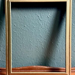 May include: A rectangular picture frame with a metallic gold finish. The frame features a stepped, tiered profile with clean, straight edges and mitered corners. The surface of the gold molding shows subtle signs of wear along the inner and outer edges.