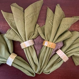 May include: Olive green cloth napkins, fanned out and held together with decorative napkin rings. The napkins have a stitched design along the edges. The napkin rings are in shades of cream, tan, and yellow.