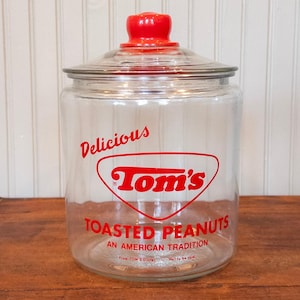 May include: A clear glass jar with a bright red lid featuring the text Delicious Tom's TOASTED PEANUTS AN AMERICAN TRADITION in red lettering. The jar has a wide cylindrical body and a rounded glass lid with a red knob handle on top.
