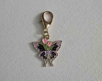 May include: A gold-colored metal butterfly charm with a lobster clasp. The butterfly has purple and black wings with pink flower accents. The charm is a decorative accessory.