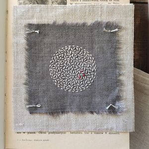 Puede incluir: Un cuadrado de tela gris con un diseño circular bordado en blanco y un pequeño acento rojo, sujeto a una tela beige. La tela está colocada sobre un libro abierto con texto en polaco.