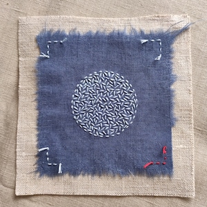 Puede incluir: Un parche textil cuadrado con un centro azul y un borde beige. El centro azul presenta un círculo bordado blanco. Las costuras blancas y rojas acentúan las esquinas. Los bordes de la tela azul están deshilachados.