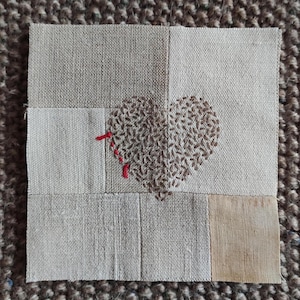 Può includere: Un tessuto patchwork quadrato con un motivo a cuore. Il cuore è ricamato con filo marrone, su uno sfondo di quadrati di tessuto beige e marrone chiaro. Accenti di filo rosso bordano il cuore.