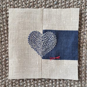 Puede incluir: Un trozo de tela cuadrado con un diseño de corazón cosido. El corazón está dividido, con una mitad en azul y la otra en blanco. El fondo es de lino natural, con una franja azul oscuro y un pequeño acento rojo.