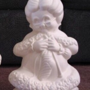 May include: A white ceramic figurine of a seated woman knitting. The figure wears glasses and has a detailed hairstyle. The figurine has a textured, decorative trim around the bottom and sleeves.