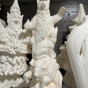 May include: Unpainted ceramic figurines of a bear standing atop a bison, with a tree-shaped figure to the left. The bear has its arm raised. The figurines are white and appear ready for painting.