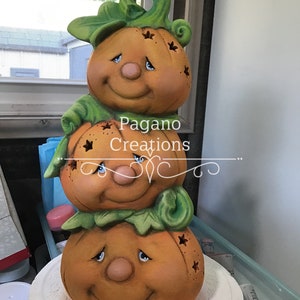 May include: A stacked pumpkin figurine with three smiling faces, each with blue eyes and a rosy nose. The pumpkins are orange with green leafy accents. The text "Pagano Creations" is visible on the middle pumpkin.