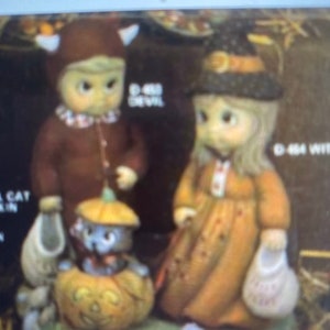 May include: A ceramic Halloween figurine set featuring a devil, a witch, and a cat in a pumpkin. The devil is in a brown costume with white horns. The witch wears a black hat and orange dress. The cat sits in a carved pumpkin.