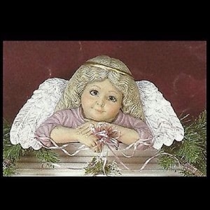 May include: A ceramic angel figurine with a serene expression, blonde curly hair, and a gold halo. The angel wears a pink top and has large white feathered wings. The figurine is holding a pink ribbon and is decorated with greenery.