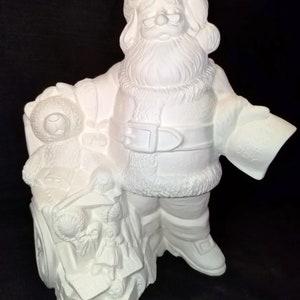 May include: A white ceramic Santa Claus figurine, holding a list and a toy-filled sleigh. The Santa figure is wearing a hat, glasses, and a belt. The sleigh is filled with toys and has wheels. The word "JOY" is visible on the list.