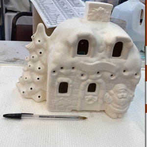 May include: Unpainted ceramic Christmas village house with a chimney, windows, and a Santa Claus figure. The house has a snow-covered roof and a Christmas tree on the side. A pen is placed next to the house for scale.