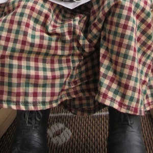 May include: A person wearing a long plaid skirt in red, green, and tan. The person is wearing black leather boots. A ball of gray yarn sits on the person's lap.