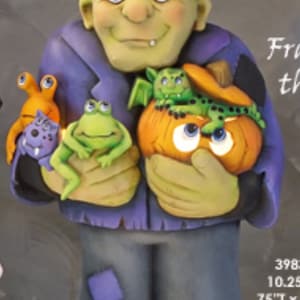 May include: A whimsical Frankenstein's monster figurine holding a pumpkin and other small creatures. The figure is painted in shades of green, purple, and gray, with a height of 17 inches and a width of 10.25 inches. The base reads "Cauldron Box".