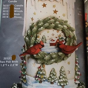 May include: A winter-themed candle with inset details. The candle features a white wax design with a melting top, gold stars, and a wreath with two red birds. The base includes snow and miniature Christmas trees.