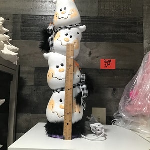 May include: A stacked Halloween decoration featuring four white ghost figures with smiling faces and orange accents. Each ghost has a black and white checkered scarf and black fur trim. The decoration is approximately 12 inches tall.