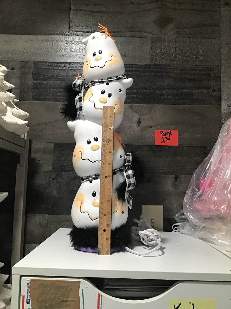 Pode incluir: Decora&ccedil;&atilde;o empilhada de Halloween com quatro figuras de fantasmas brancos com rostos sorridentes e detalhes em laranja. Cada fantasma tem um cachecol xadrez preto e branco e acabamento em pele preta. A decora&ccedil;&atilde;o tem cerca de 30 cm de altura.