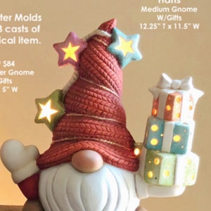 May include: A ceramic gnome figurine with a red knit hat adorned with glowing star accents. The gnome has a white beard, a tan nose, and is holding a stack of illuminated gift boxes. The text on the image reads "Hans Medium Gnome W/Gifts 12.25" T x 11.5" W".
