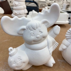 May include: A white ceramic moose figurine with large antlers, a scarf, and a smiling face, holding a round ornament. The moose is part of a collection of similar unpainted ceramic figurines, perfect for DIY painting projects and holiday decorations.
