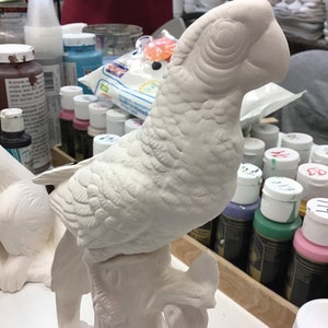 May include: A detailed, unpainted ceramic parrot figurine perched on a tree branch. The bird has textured feathers and a curved beak. The figurine is ready to be painted and is a craft item.
