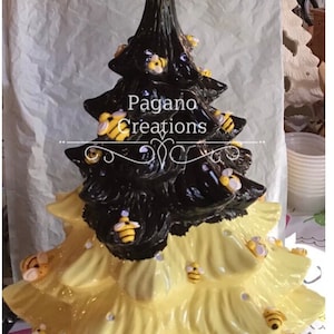 May include: A ceramic Christmas tree decorated with bees and the words "Pagano Creations" and "Bee Kind". The tree is painted black and yellow and has a wooden stick with a beehive on top.
