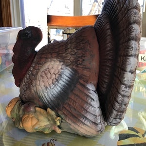 May include: A ceramic turkey figurine with brown, white, and red coloring. The turkey has a detailed tail and is resting on a base with autumn leaves and pumpkins. The figurine is likely a decorative item for the Thanksgiving holiday.
