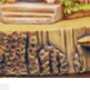 May include: A decorative miniature fairy house on a tree stump base. The base is a light brown color with a textured bark design. A small orange mushroom with white spots is to the right of the base. The fairy house has a light blue door and a green roof.