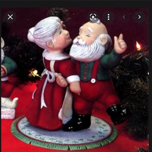 May include: A ceramic Christmas figurine set featuring Santa Claus and Mrs. Claus. Santa wears a red suit with green suspenders, while Mrs. Claus is in a red dress. They are standing on a round rug with a green border, with a festive background.