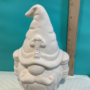May include: A white ceramic gnome figurine with a tall, pointed hat featuring a decorative emblem on the front and a full beard. A wooden ruler is positioned vertically next to the gnome, showing a height of approximately 8 inches tall.