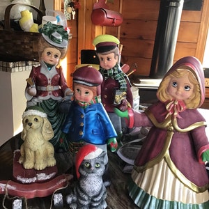 May include: A collection of five hand-painted ceramic figurines, including a girl in a red dress, a boy in a blue coat, a girl in a pink dress, a dog, and a cat. The figurines are all wearing festive attire and are arranged on a marble surface.