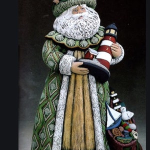 May include: A ceramic Santa Claus figurine, dressed in a green and gold coat with white trim. He holds a red and white striped lighthouse and a small house. A sack filled with toys and a sailboat is at his feet.