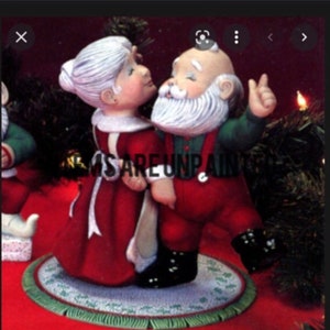May include: A ceramic figurine set of Mr. and Mrs. Claus in a festive pose. Mrs. Claus wears a red dress with white trim, while Mr. Claus sports a red suit and green shirt. The figures stand on a round, green-trimmed mat.