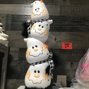 May include: A stacked Halloween decoration featuring three white ghost figures with smiling faces, black and white plaid scarves, and black feather trim. Orange accents and star cutouts add detail to this festive, light-up decor.