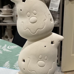 May include: A stack of four unpainted ceramic ghost figures. Each ghost has a smiling face, star-shaped cutouts, and a paw print design. The figures are stacked on top of each other, creating a whimsical decorative piece.