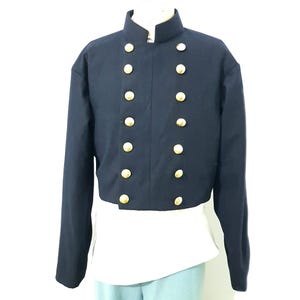 Könnte beinhalten: Marineblaue Jacke mit Stehkragen und goldfarbenen Knöpfen. Die Jacke hat lange Ärmel und einen doppelreihigen Schnitt. Sie wird über einem weißen Hemd und hellblauen Hosen getragen.