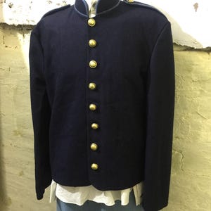 Könnte beinhalten: Marineblaue Jacke mit Stehkragen und goldenen Knöpfen. Die Jacke hat eine taillierte Passform und wird über einem weißen Hemd und einer blauen Hose getragen. Das Design der Jacke erinnert an historische Militäruniformen.