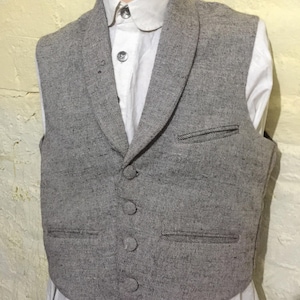 May include: A grey tweed waistcoat with a shawl collar and four buttons. The waistcoat has two front pockets and is worn over a white collared shirt. A classic style.