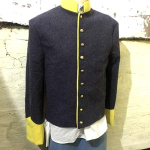 May include: A navy blue jacket with a yellow collar and cuffs. The jacket has a row of gold buttons down the front and is worn over a white shirt and blue pants. The jacket appears to be made of wool.